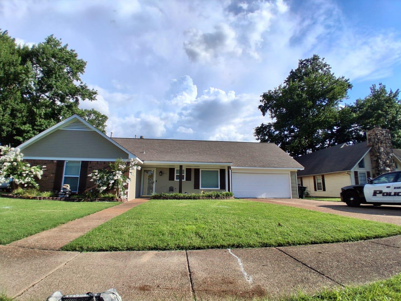 A single-story suburban house with a neatly mowed lawn, white flowers, and a police car parked in the driveway on a sunny day.