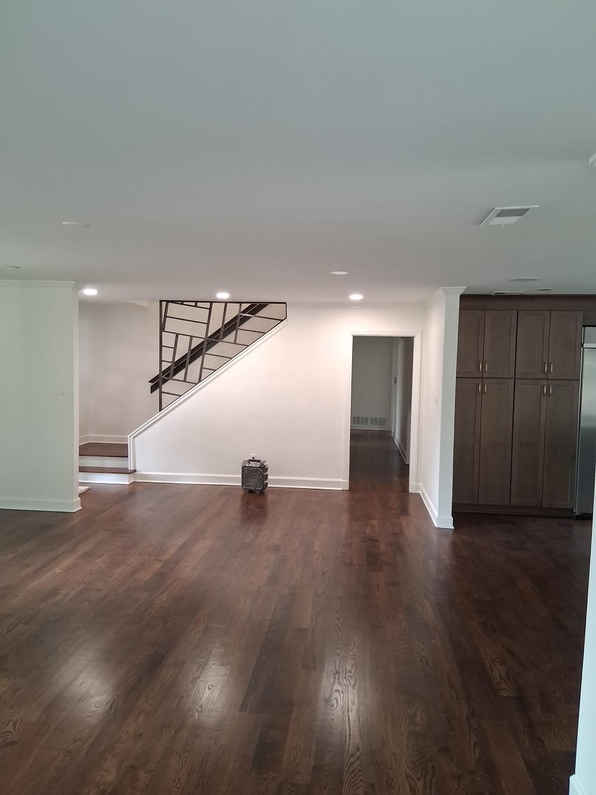 An empty room with dark hardwood floors, white walls, a staircase with metal railings, built-in cabinets, and a small appliance on the floor.