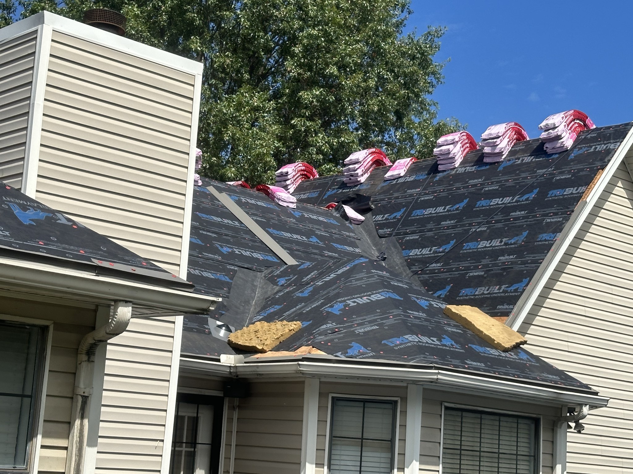 Stacks of roofing shingles sit on a house roof under renovation, with black synthetic underlayment partially installed and a blue sky in the background.