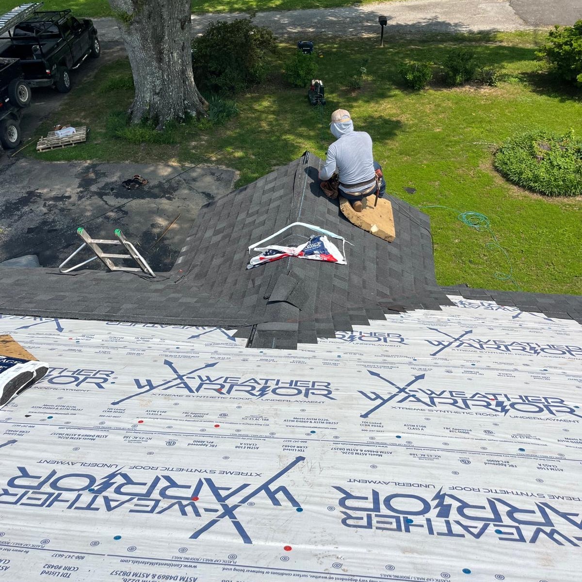 A person sits on the peak of a house roof installing shingles, with roofing underlayment labeled Weather Warrior visible beneath. Tools and materials are scattered nearby.
